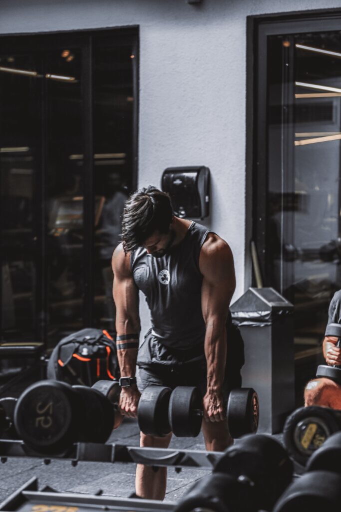 A muscular man in a tank top lifts heavy dumbbells in a gym setting.